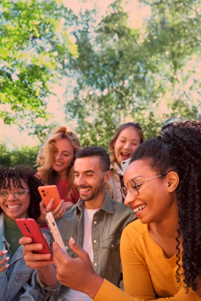 Five people of mixed races holding smartphones and engaged on social media smiling in a park with trees in the background