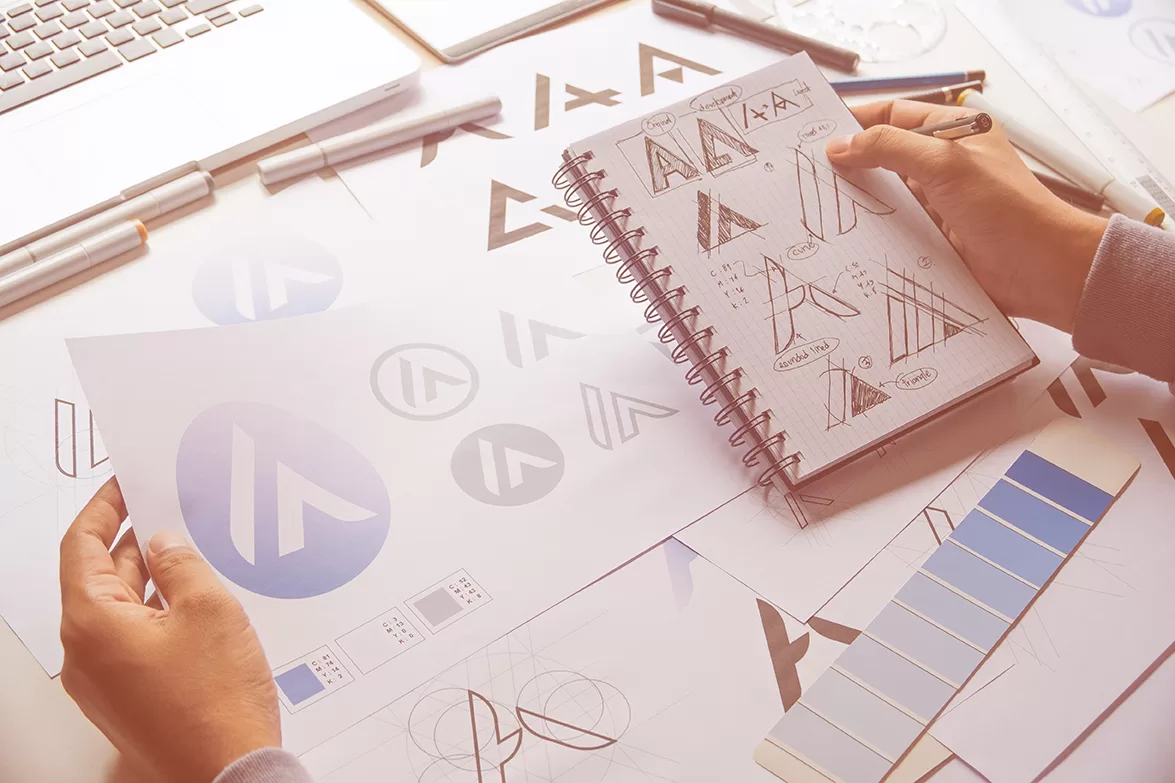 A logo designer holding a notebook with logo design sketches, surrounded by papers with logo designs, pens, and color palettes on a white table