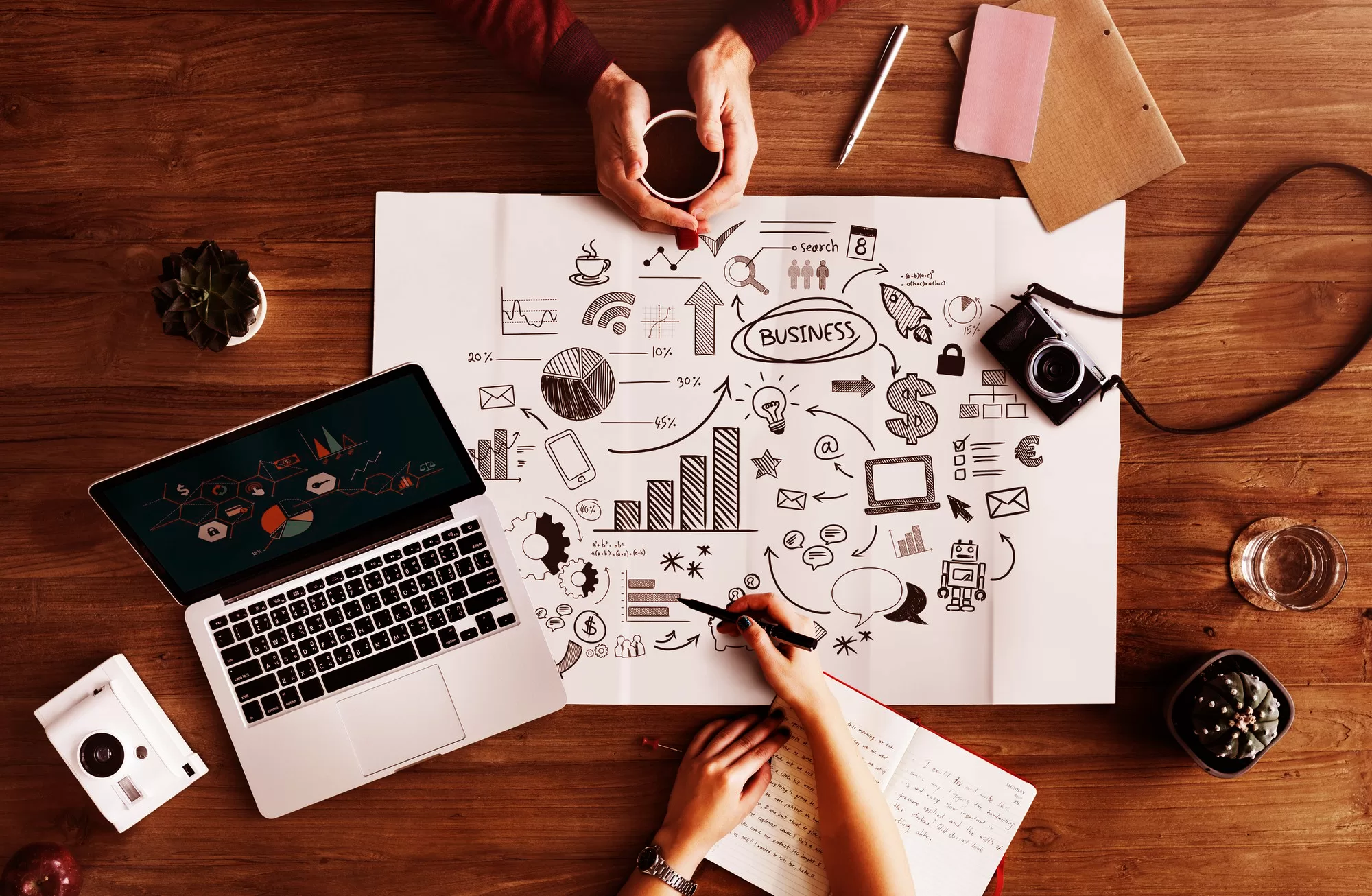 top down view of two digital marketing professioanls discussing brand and business growth diagrams on a white paper with a laptop, a camera, a notebook and a cup of coffee on a wooden table