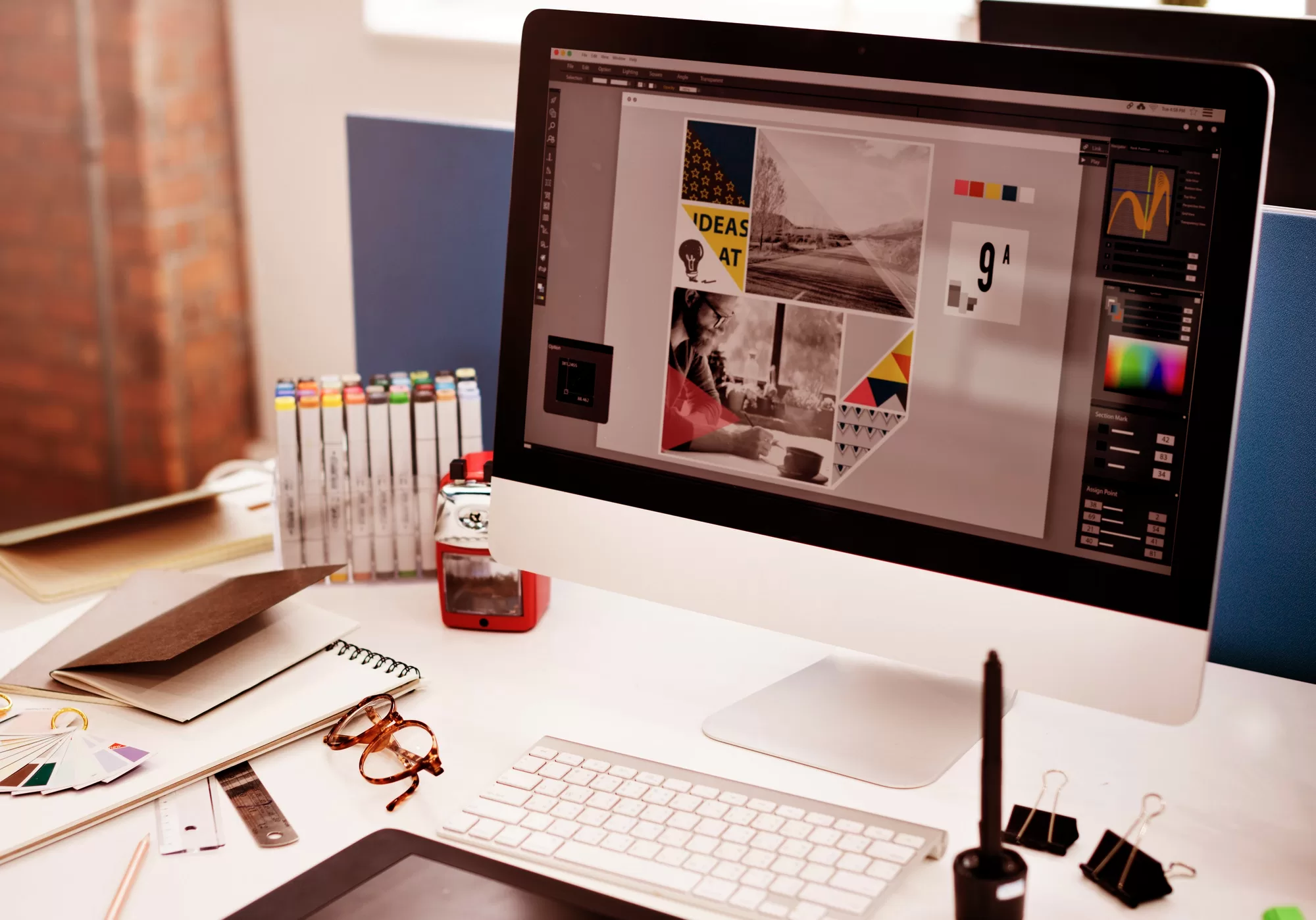 Computer screen displaying a graphic design project, keyboard, pen, glasses, markers, notebook, and other office supplies on a white desk in an office environment