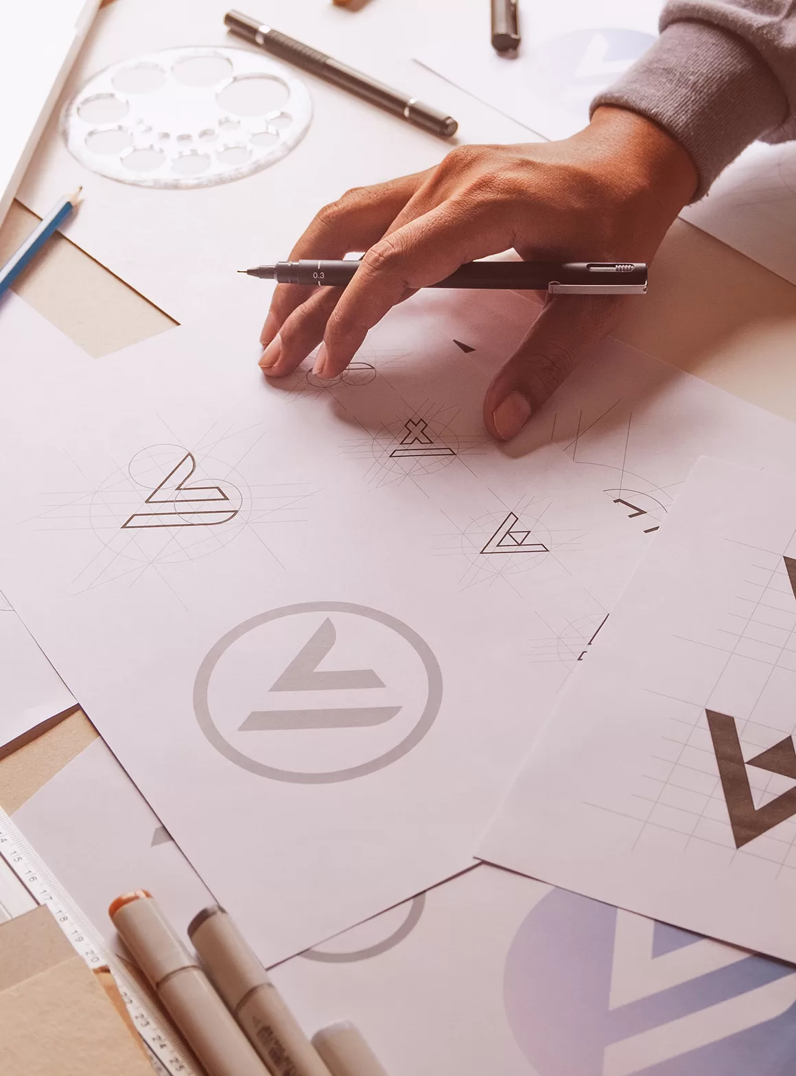 A tan-skinned graphic designer's hand holding a pen over a white paper with logo design sketches, surrounded by design tools, top-down view, bright digital marketing studio environment