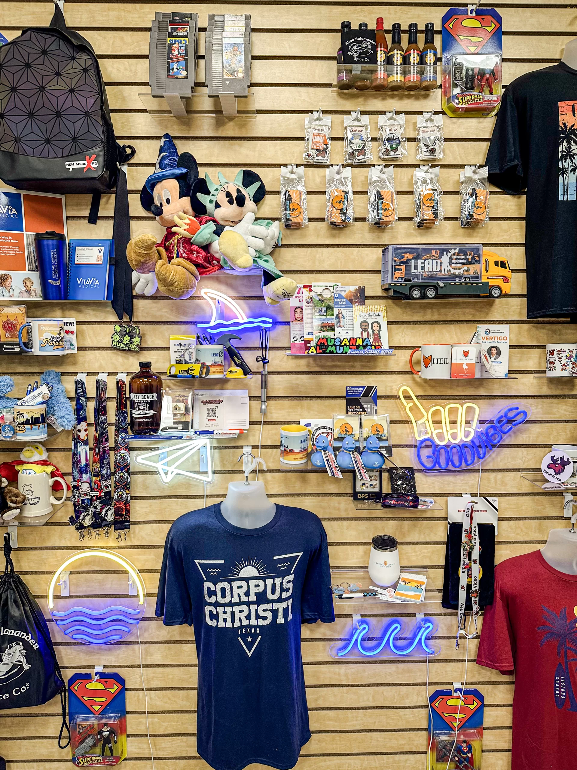 A print marketing store wall with a variety of merchandise, including plush toys, keychains, mugs, t-shirts, and neon signs of client brand logos, the background is a wooden slat wall