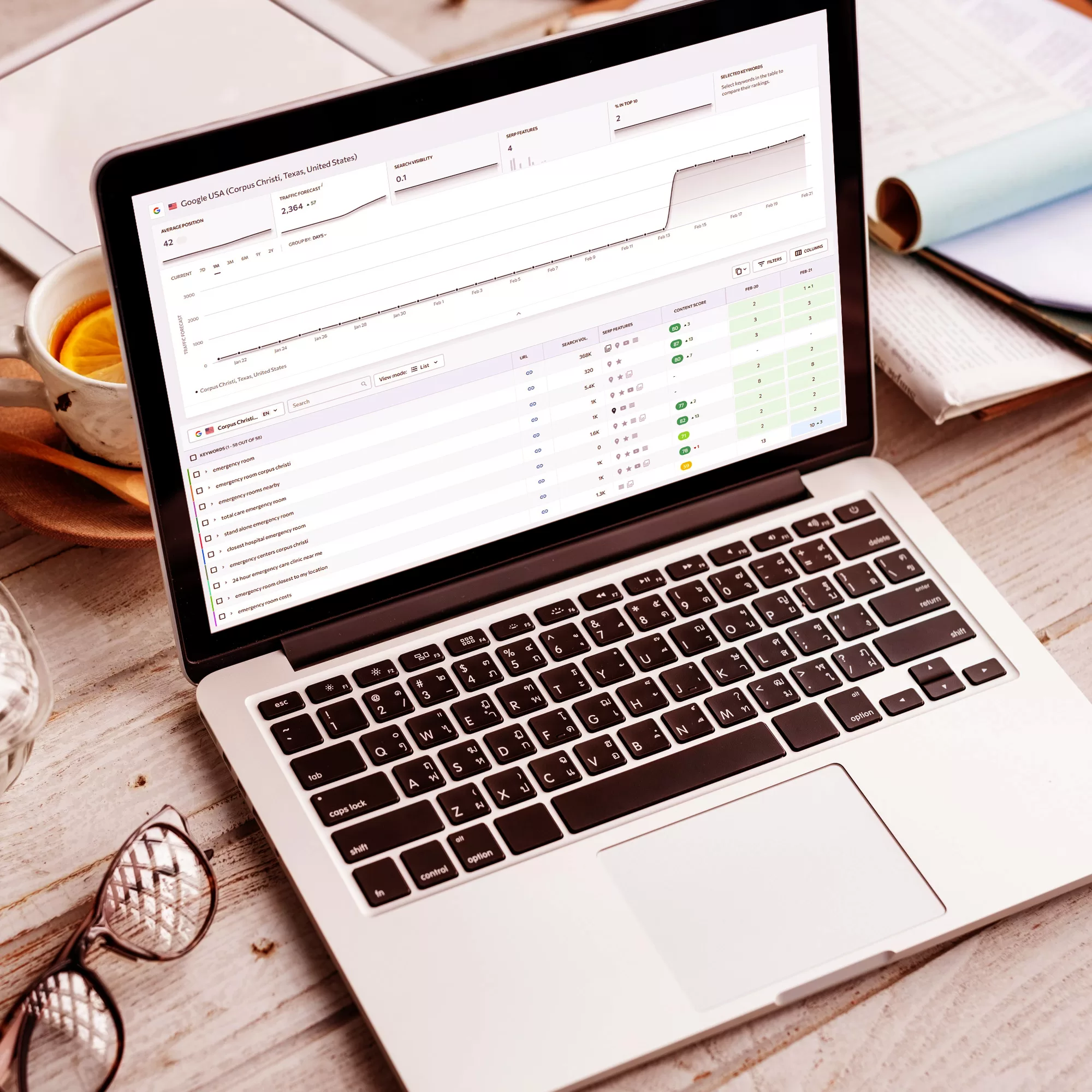 a laptop displaying a search engine optimization (SEO) rankings and data on a wooden desk, with a cup of tea, glasses, and papers scattered around